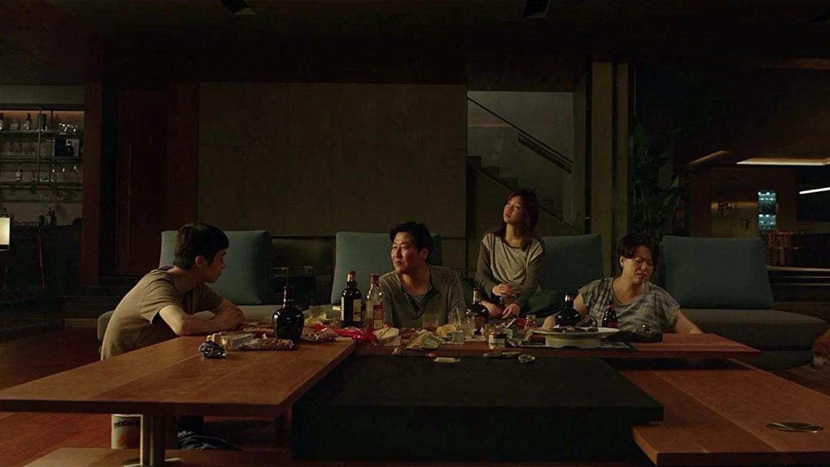 A still from Parasite showing four family members sprawled around a low wooden coffee table in a dimly lit, upscale living room, surrounded by empty liquor bottles and half-eaten snacks after a night of drinking.