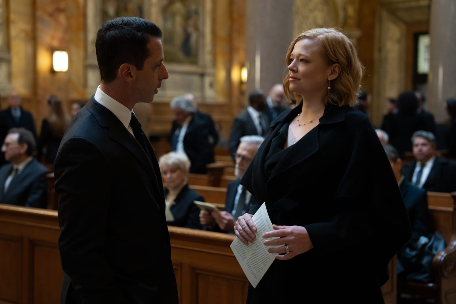 A still from Succession showing Kendall and Shiv Roy in black funeral attire, standing face to face in the aisle of an ornate cathedral, with mourners seated in pews and gold architectural details in the background.