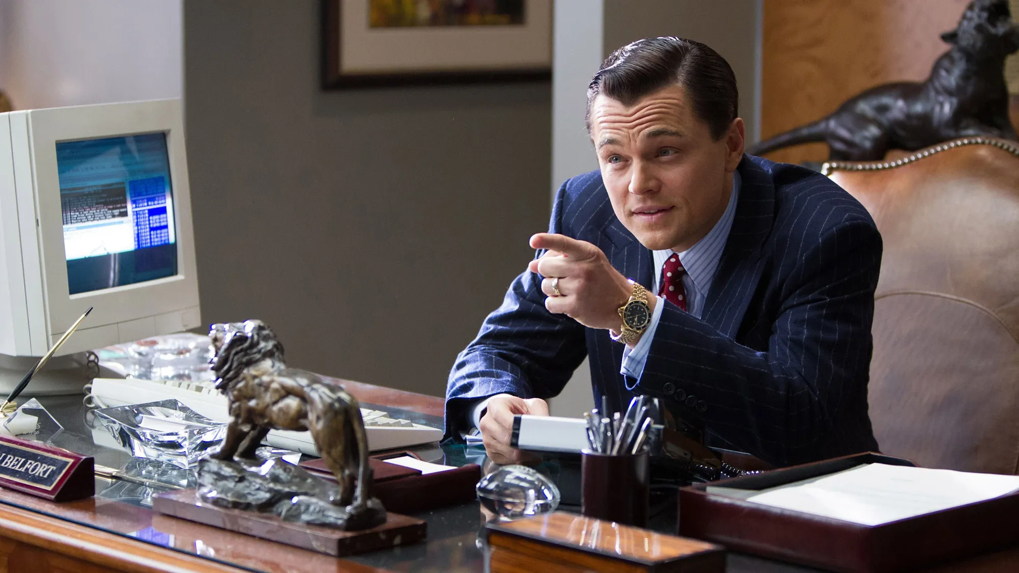 Leonardo DiCaprio as Jordan Belfort in The Wolf of Wall Street, sitting at a wood-panelled office desk in a pinstriped suit, pointing and mid-sentence, with a bronze lion figurine and vintage computer monitor on the desk in front of him.